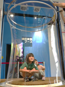 子どもたちが大きなシャボン玉の中に入る実験装置を体験している写真